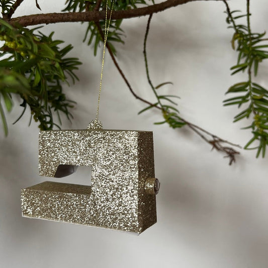 Silver glittery sewing machine ornament hanging on a branch with green leaves.