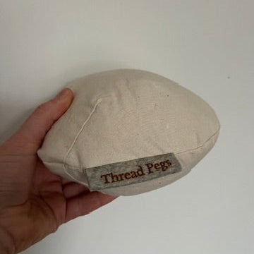 Hand holding a beige fabric tailors ham with 'Thread Pegs' label against a white background