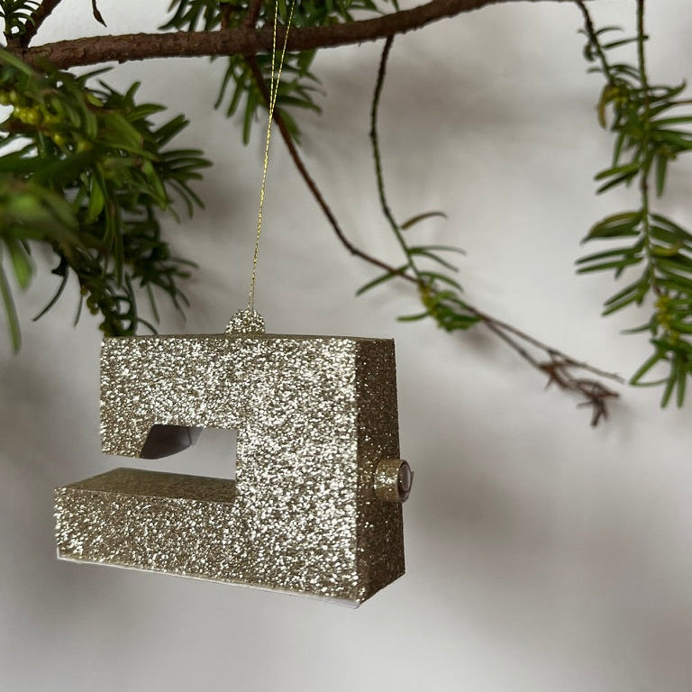 Silver glittery sewing machine ornament hanging on a branch with green leaves.